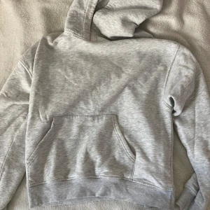 Grå hoodie från Gina Tricot - En basic ljusgrå hoodie från Gina Tricot, basically basics-kollektionen. Tröjan har huva, magficka och ribbade muddar. Perfekt för chill dagar och enkel att matcha med jeans eller mjukisbyxor.