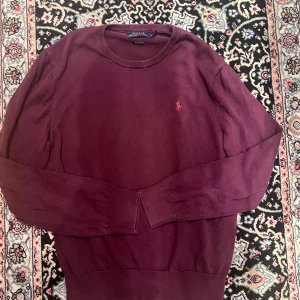 Vinröd tröja från Polo Ralph Lauren - Vinröd långärmad tröja från Polo Ralph Lauren i 100% pima bomull. Klassisk rund halsringning och broderad logga på bröstet. Perfekt för dig som gillar stilrena och tidlösa plagg med en touch av lyx.