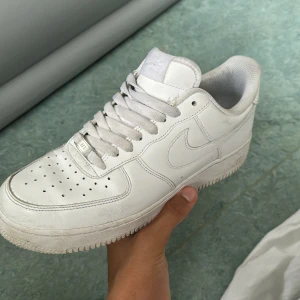 Nike Air Force 1 vita sneakers - Klassiska Nike Air Force 1 sneakers i helvitt skinn med perforerad tå, rund tå och platt sula. Snygg och tidlös modell med snörning och diskret Nike-logga på sidan och hälen. Perfekta för dig som gillar clean och stilren streetwear.