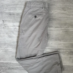 Grå chinos från Tommy Hilfiger - Snygga grå chinos från Tommy Hilfiger med klassisk passform. Byxorna har bälteshällor, sidofickor och en bakficka med knapp. Tillverkade i bomull med en diskret Tommy-detalj vid fickan. 