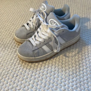 Adidas sneakers i ljusgrå färg - Snygga Adidas sneakers i ljusgrå  med klassiska vita ränder och vit sula. Skorna har rund tå, platt gummisula och vita snören. Perfekta för dig som gillar retrostil och vill ha något som sticker ut lite extra. Ny pris 2000