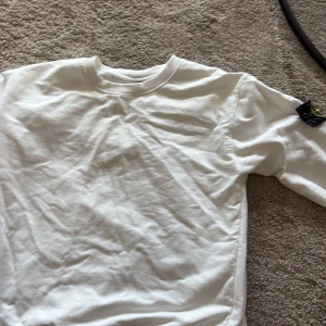 Vit långärmad tröja från Stone Island - Vit långärmad tröja från Stone Island med klassisk rund halsringning och ikoniskt svart märke med gul och grön brodyr på vänster ärm. Tröjan är i mjuk bomull och har en clean, stilren look som passar perfekt till jeans eller joggers.