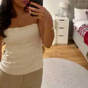 Vit korsett topp i satin-look - Säljer en snygg vit korsett topp från Zara med ”satin-look”. Toppen är axelbandslös och har rynkade detaljer samt hakknäppning bak. Perfekt för dig som vill ha en clean och stilren vibe till outfiten. Går att styla på många sätt! Det är lite sönder där bak (se bild 4) men inget som syns när den är helt knäppt.