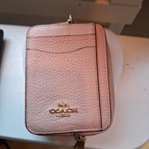 Ljusrosa plånbok från Coach i skinn - Supersöt ljusrosa plånbok från Coach i äkta skinn med guldiga detaljer. Plånboken har flera kortfack, dragkedja och ett fönster för ID eller foto på baksidan. Perfekt storlek för att ha med sig överallt och snygg att matcha med andra accessoarer.