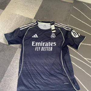 Real Madrid Adidas fotbollströja M - Snygg Real Madrid fotbollströja från Adidas i marinblått med tunna ränder och vita detaljer. Tröjan har korta ärmar, broderad klubbmärke och Adidas-logga samt sponsortryck på bröstet. Tillverkad i lätt och ventilerande material, perfekt för match eller träning.