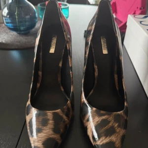 Leopardmönstrade pumps från Guess - Snygga pumps från Guess med leopardmönster i bruna och svarta toner. Skorna har spetsig tå, hög klack och en glansig finish. På baksidan finns en liten metallogo för extra detalj. Perfekta för dig som vill sticka ut med din stil.