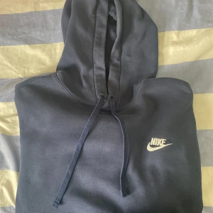 Mörkblå Nike hoodie med broderad logo - Säljer en mörkblå hoodie från Nike i storlek L. Tröjan har klassisk huva med snörning och en snygg broderad Nike-logga på bröstet. Perfekt för dig som gillar en enkel och stilren look. Materialet är mjukt och skönt, troligtvis bomullsmix.