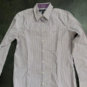 Lila och vit randig skjorta från GANT - Snygg skjorta från GANT i lila och vitt randigt mönster. Klassisk krage, långa ärmar och knäppning framtill med vita knappar. Broderad GANT-logga på bröstet. Skjortan är i bomull och har en stilren, tidlös look. Knappast använd, vill bara bli av med 