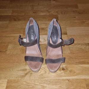 Armani Jeans sandaler med klack - Snygga sandaler från Armani Jeans med hög klack och öppen tå. Skorna har grått glittrigt tyg på hälen, bruna remmar i skinn och en stadig träfärgad klack. Perfekta för dig som vill sticka ut med en cool och elegant look.