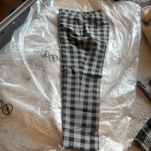 Svarta och grå rutiga kostymbyxor - Snygga kostymbyxor i svart och grått med rutigt mönster. Byxorna har raka ben, bälteshällor och knapp bakficka. Perfekta för dig som vill sticka ut med en klassisk men trendig look. Midjan är 30 tum och benlängden 30 tum.