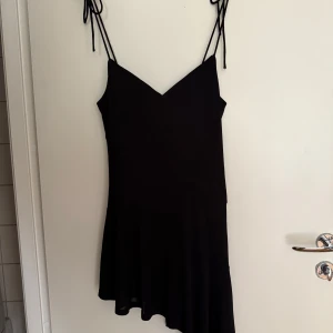 Svart kort klänning med smala axelband - Supersnygg svart kort klänning med smala knytbara axelband och v-ringad hals. Klänningen har en lätt utsvängd nederdel och asymmetrisk fåll som ger en cool och trendig look. Perfekt för dig som gillar minimalistisk stil och vill ha något enkelt men ändå stilrent.
