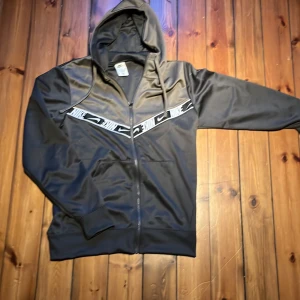Svart grå nike zip upp med huva - Svart grå zip upp från Nike med hel dragkedja, huva och vita detaljer med Nike-loggor över bröstet. Jackan har två fickor framtill, perfekt för en sportig look. Snygg och enkel att matcha till jeans eller träningsbyxor.
