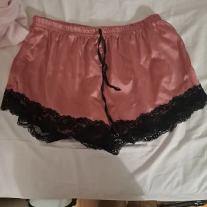 Rosa satinsleep-shorts med spets - Snygga rosa shorts i satin-look med svart spetskant nertill och snörning i midjan. De har en loose fit och känns riktigt mjuka och lyxiga, perfekta för chill eller som pyjamasshorts. Färgen är dämpad rosa och materialet är glansigt.