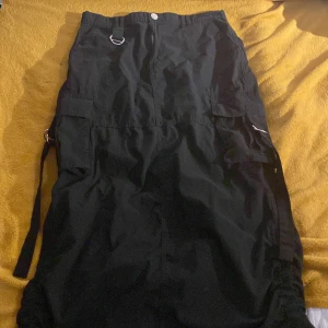 Svarta cargokjol med detaljer - Svarta cargokjol med flera fickor, D-ringar och justerbara band i sidorna. Byxorna har en loose passform och är tillverkade i ett slitstarkt bomullstyg. Perfekta för dig som gillar streetwear och funktionella detaljer.