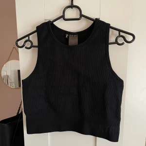 Svart ribbad tränings topp från Stronger - Snygg svart ribbad tränings topp från Stronger med croppad passform. Toppen är ärmlös och har breda axelband samt diskret logga nertill. Perfekt för träning eller chill dagar. Materialet är stretchigt och följsamt för maximal komfort. Jag är osäker på storlek då det inte står i toppen men jag gissar att det är en M. (Porto tillkommer) 