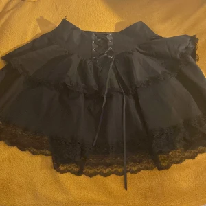 Svart volangkjol med snörning - Supersöt svart kortkjol med två lager volanger och spetsdetaljer längst ner. Hög midja med snygg snörning framtill som ger en cool touch. Kjolen är fluffig och har dragkedja bak. Perfekt för dig som gillar alternativ eller gothstil.