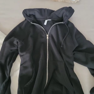 Svart hoodie med dragkedja 💗 - Svart hoodie från Divided i storlek XXS. Klassisk modell med huva, snörning och hel dragkedja framtill. Mjuk bomullsblandning och två fickor framtill. Perfekt för chill dagar eller när du vill ha en enkel och stilren look.