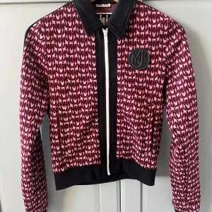Miu Miu mönstrad jacka - Snygg bomberjacka från Miu Miu med coolt röd, svart och vit mönster över hela jackan. Jackan har svart krage, muddar och dragkedja framtill samt ett stort Miu Miu-emblem på bröstet. Perfekt statement-plagg för dig som vill sticka ut. Står som M men passar som en XS.
