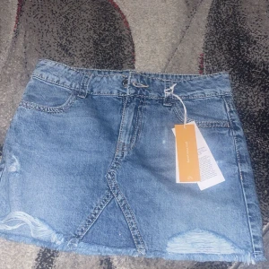 Blå jeanskjol med rå kant från Kappahl - Snygg blå jeanskjol från Gina i klassisk modell med rå kant nertill och slitna detaljer. Kjolen har framfickor, bälteshällor och två bakfickor med knapp. Perfekt för dig som gillar en trendig och avslappnad stil. 