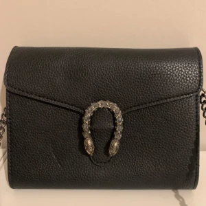 Svart axelväska med kedja och ormdetalj - Snygg svart axelväska i skinn med cool kedjerem och unik ormdetalj i silver på framsidan. Väskan har flera fack och dragkedja inuti, perfekt för att hålla ordning på dina grejer. Stilren och edgy design som lyfter vilken outfit som helst.