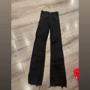 Svarta jeans med slits och rå kant - Stretchiga. Säljer ett par svarta jeans med raka ben och slits nertill. Byxorna har råa, fransiga kanter längst ner för en edgy look. Klassisk femficksmodell med knapp och dragkedja. Perfekta till sneakers eller boots.
