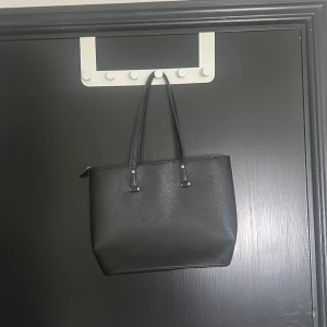 Svart axelväska i skinn - Stilren svart axelväska med två långa handtag och enkel design. Väskan är rymlig och har dragkedja upptill. Perfekt nu till skolstarten!            OBS! Den ser större ut än vad den är i verkligheten.  De står inget märke på den, men den är köpt på Gekås Ullared.