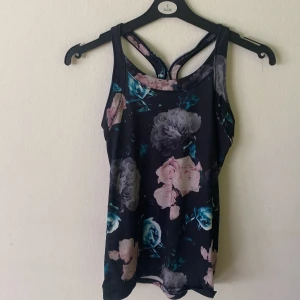 Blommig träningstopp från Phelix Sports - Supersnygg svart träningstopp från Phelix Sports med blommigt mönster i rosa, grått och blått. Modellen är ärmlös med brottarrygg och sitter tight mot kroppen. Perfekt för gymmet eller löprundan, tillverkad i stretchigt syntetmaterial som andas.