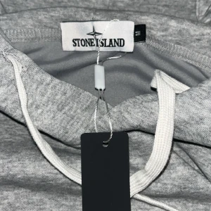 Grå hoodie från Stone Island - Aldrig använd. Snygg grå hoodie från Stone Island med klassisk logga på ärmen. Tröjan har huva med vita snören och en stor magficka. Perfekt för dig som gillar streetwear och vill ha en stilren look med premiumkänsla. 1 av 1 