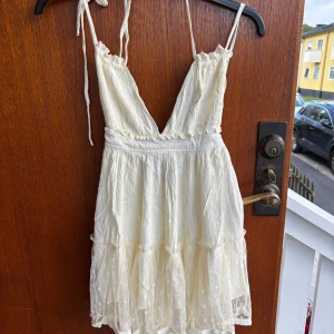 Vit kort sommarklänning med volang - Superfin vit kort klänning med smala axelband och djup v-ringning. Kjolen har flera lager volang i mesh med prickigt mönster och ger en luftig känsla. Perfekt för varma dagar och har knytband både upptill och baktill för extra detalj.