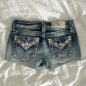 Miss Me jeansshorts med bling i strl 26 - Snygga mörkblå jeansshorts från Miss Me med blingade detaljer och fina fickor bak. Shorts med låg midja, slitningar och silverfärgade knappar med logga. Perfekta för sommaren när du vill sticka ut lite extra. Midjemått: ca 36 cm