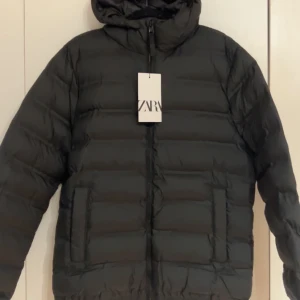 Helt ny Svart pufferjacka från Zara - Svart pufferjacka från Zara med huva och dragkedja framtill. Jackan har en quiltad look med horisontella sömmar och två fickor på sidorna. Perfekt för kalla dagar när du vill hålla stilen enkel och clean.