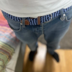 Blåvitt mönstrat bälte  - Säljer ett snyggt bälte med blåvitt zickzackmönster och bruna skinn-detaljer vid spännet. Bältet är elastiskt och passar perfekt till jeans för en cool och avslappnad look. Helt nytt!