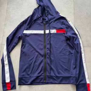 Blå vindjacka med ränder från SHEIN - Sportig blå vindjacka med huva, hel dragkedja och vita samt röda detaljer längs ärmar och bröst. Jackan har en clean look med smala ränder och är perfekt för en chill streetstyle. Materialet är lätt och känns skönt mot huden.