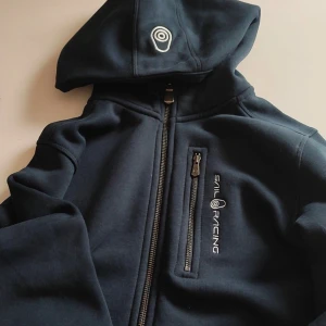 Marinblå Sail Racing hoodiejacka - Mörkblå hoodiejacka från Sail Racing med dragkedja och bröstficka med zip. Jackan har broderad logga på bröstet och huvan, samt en sportig och clean look. Perfekt för dig som gillar stilren design och funktionella detaljer.
