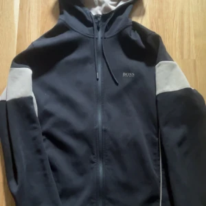 Hugo boss  - Svart hoodie från Hugo Boss med gråa detaljer på ärmarna och insidan av huvan. Tröjan har hel dragkedja framtill, snörning i huvan och diskret Boss-logga på bröstet. Perfekt för en chill och stilren look.