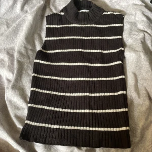 Randig linne med en krage  - Snygg stickad väst i mörkbrunt med vita horisontella ränder från Gina Tricot. Modellen är ärmlös med hög krage och ribbad struktur, perfekt att styla över en skjorta eller t-shirt för en trendig lager-på-lager-look.