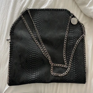 Svart axelväska Stella McCartney - Säljer en svart axelväska från Stella McCartney med coolt ormskinnsmönster och silverfärgad kedja. Väskan har en stor rund logga i metall och rymligt innerfack med dragkedja. Perfekt för dig som vill ha en statementväska med edgy vibe. Priset går att diskutera eller om du vill byta den mår något annat 