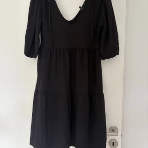 Svart klänning från TSI Melissa S - Svart klänning från TSI i modellen Melissa. Klänningen har en djup v-ringning både fram och bak, trekvartsärmar och en luftig, voluminös passform med flera volanger. Materialet är lätt strukturerat och känns bekvämt mot huden. Strl S helt oanvänd pga fel storlek 