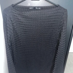 Svart långärmad tröja från 157 - Svart långärmad tröja från 157 med vågig struktur i tyget. Tröjan har en klassisk rund halsringning och är tillverkad i ett mjukt, stretchigt material som känns skönt mot huden. Perfekt för lager-på-lager och enkel att matcha med olika outfits.