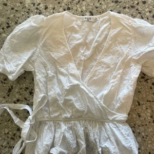 Vit omlottblus från NA-KD - Superfin vit omlottblus från NA-KD med broderade blommor, puffiga korta ärmar och knyt i midjan. Blusen har v-ringning och är gjord i bomull, perfekt för sommarens alla tillfällen.