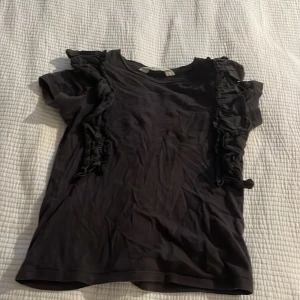 Svart volangblus från H&M - Svart blus från H&M med korta ärmar och snygga volangdetaljer längs axlarna. Toppens material är mjukt och lätt, perfekt för att ge outfiten en extra touch. Enkel att matcha med både jeans och kjol för en trendig look.