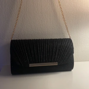 Svart glittrig kuvertväska med kedja - Snygg svart festväska med glittrigt tyg och veckad design. Väskan har en lång guldfärgad kedja. Man kan antingen ha kedjan kort på axeln eller så kan man förlänga kedjan och ha den på ett annat sätt! 