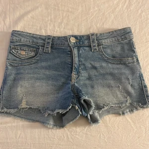 Blå jeansshorts med fransig kant - Snygga blå jeansshorts med fransig kant och slitna detaljer. Klassisk femficksmodell med knapp och dragkedja framtill samt coola bakfickor med knapp. Perfekta för varma dagar och en avslappnad stil.