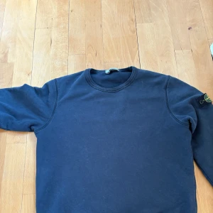 Mörkblå sweatshirt från Stone Island - Mörkblå sweatshirt från Stone Island Junior med klassisk rund hals och ikonisk logga på vänster ärm. Tröjan har ribbade muddar och är tillverkad i mjuk bomull. Perfekt för dig som gillar streetwear och vill ha en clean look. Jag har även kvittot till tröjan.