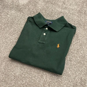 Mörkgrön pikétröja från Ralph Lauren - Snygg mörkgrön pikétröja från Polo Ralph Lauren med klassisk krage och knäppning framtill. Tröjan har korta ärmar och den ikoniska orange broderade loggan på bröstet. Perfekt för en clean och sportig look.