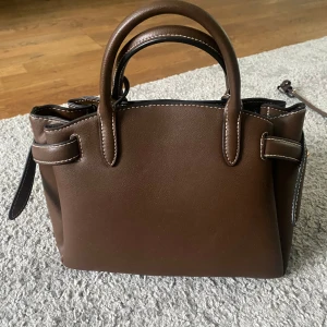 Brun handväska med vita sömmar - Säljer en snygg brun handväska med vita kontrastsömmar och dubbla handtag. Väskan har dekorativa remmar på sidorna och är perfekt för att bära dina viktigaste saker. Enkel och stilren design som passar till många outfits.