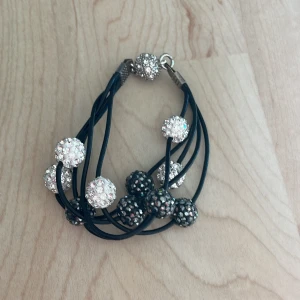 Svart armband med glittriga pärlor - Säljer ett svart armband med flera glittriga pärlor i silver och svart. Armbandet har flera svarta band och pärlorna är täckta av små gnistrande stenar. 