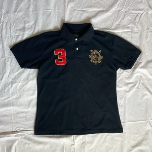 Svart pikétröja från Polo Ralph Lauren - Svart pikétröja från Polo Ralph Lauren med röd broderad siffra 3 på bröstet och stort broderat emblem både fram och bak. Klassisk krage och knäppning med två knappar. Perfekt för en sportig och stilren look. Strl xl i barn så typ S/M i vuxenstorlek. 170-175cm