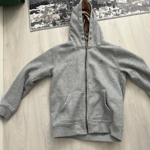 Grå hoodie med dragkedja från Burberry - Säljer en grå hoodie från Burberry med klassiskt rutigt foder i huvan och insidan. Jackan har dragkedja framtill, två stora fickor och ribbade muddar. Perfekt för dig som gillar stilrena plagg med en twist.