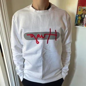 Vit sweatshirt med röd broderad Hugo Boss - Säljer en vit sweatshirt med en grå rektangel och röd broderad text på bröstet. Tröjan har rund halsringning och långa ärmar. Perfekt för en stilren och avslappnad look. Köpt för 1200kr, knappt använd, säljer för halva priset‼️